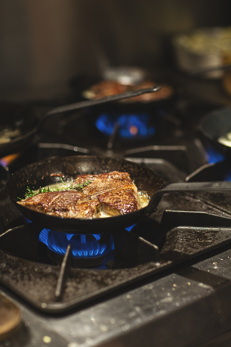 Close-up van een gietijzeren pan met aangebakken vlees en kruiden boven een blauwe gasvlam