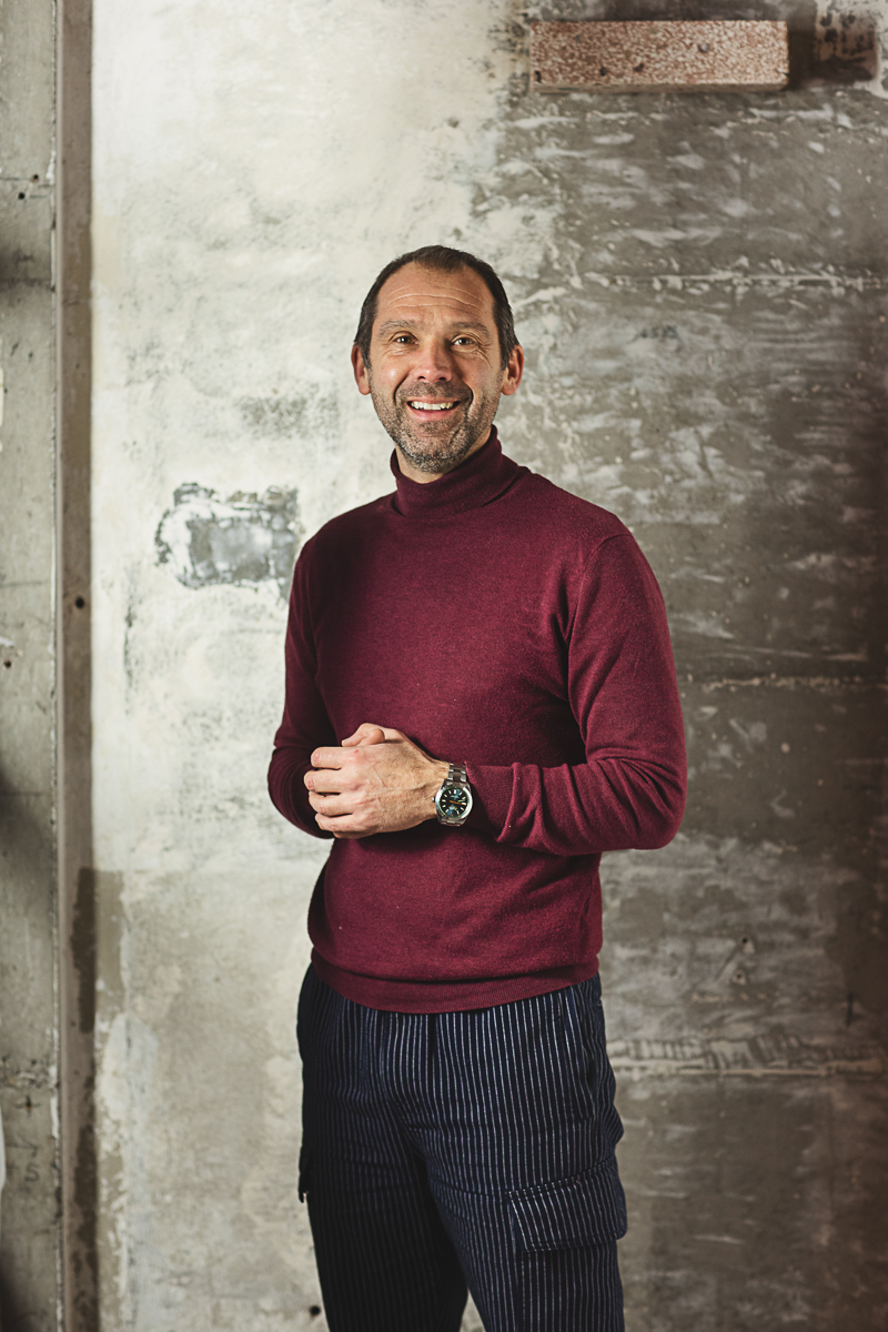 Portretfoto van Fitzgerald Fitz Rigole in bordeauxrode coltrui voor ruwe betonnen muur