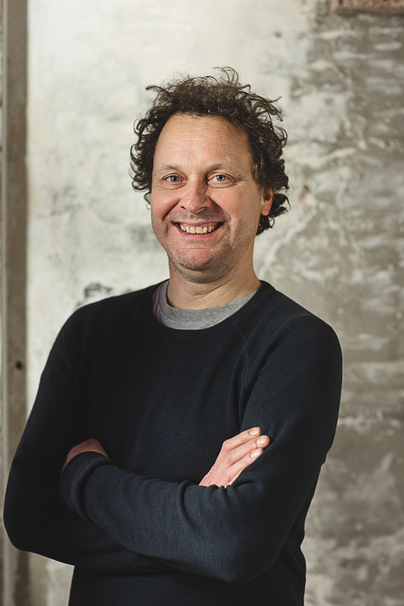 Portretfoto van Tom Brys met krullend haar en glimlach in donkere trui voor betonnen muur
